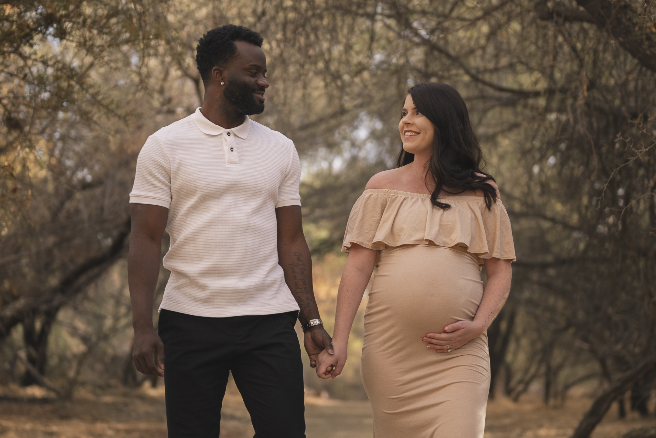 Pregnant couple walk through tunnel of trees holding hands during Arizona maternity session