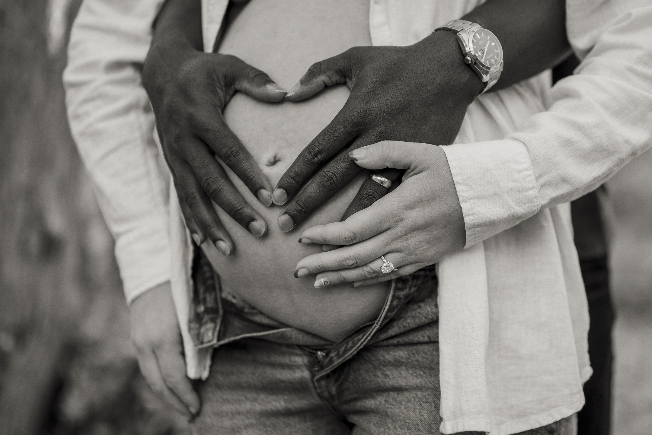 Black and white photo of father and mother's hands on pregnant belly forming a heart during maternity photo session