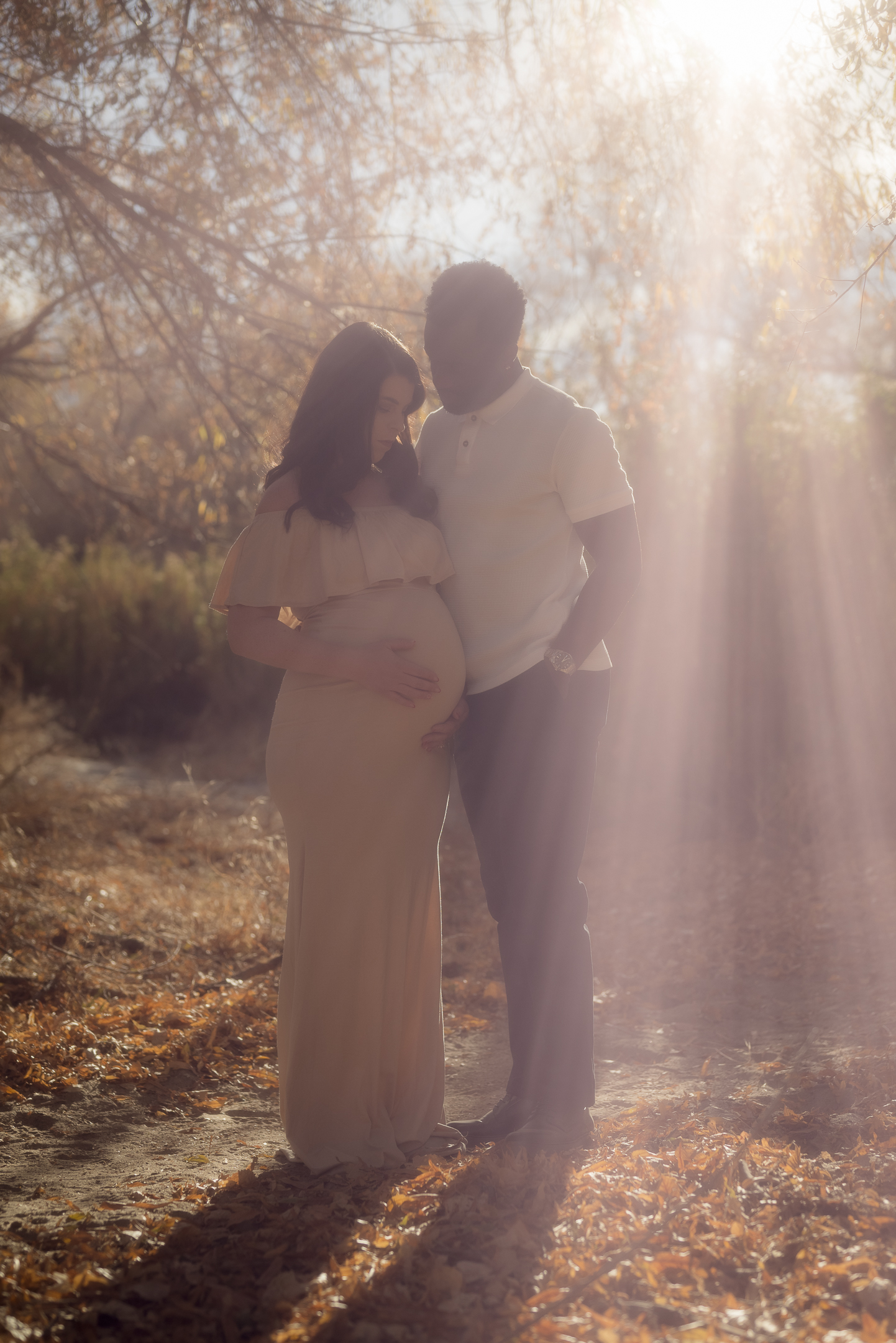 Couple standing in a dreamlike setting with rays of sunlight shining down on them as they admire her pregnant stomach