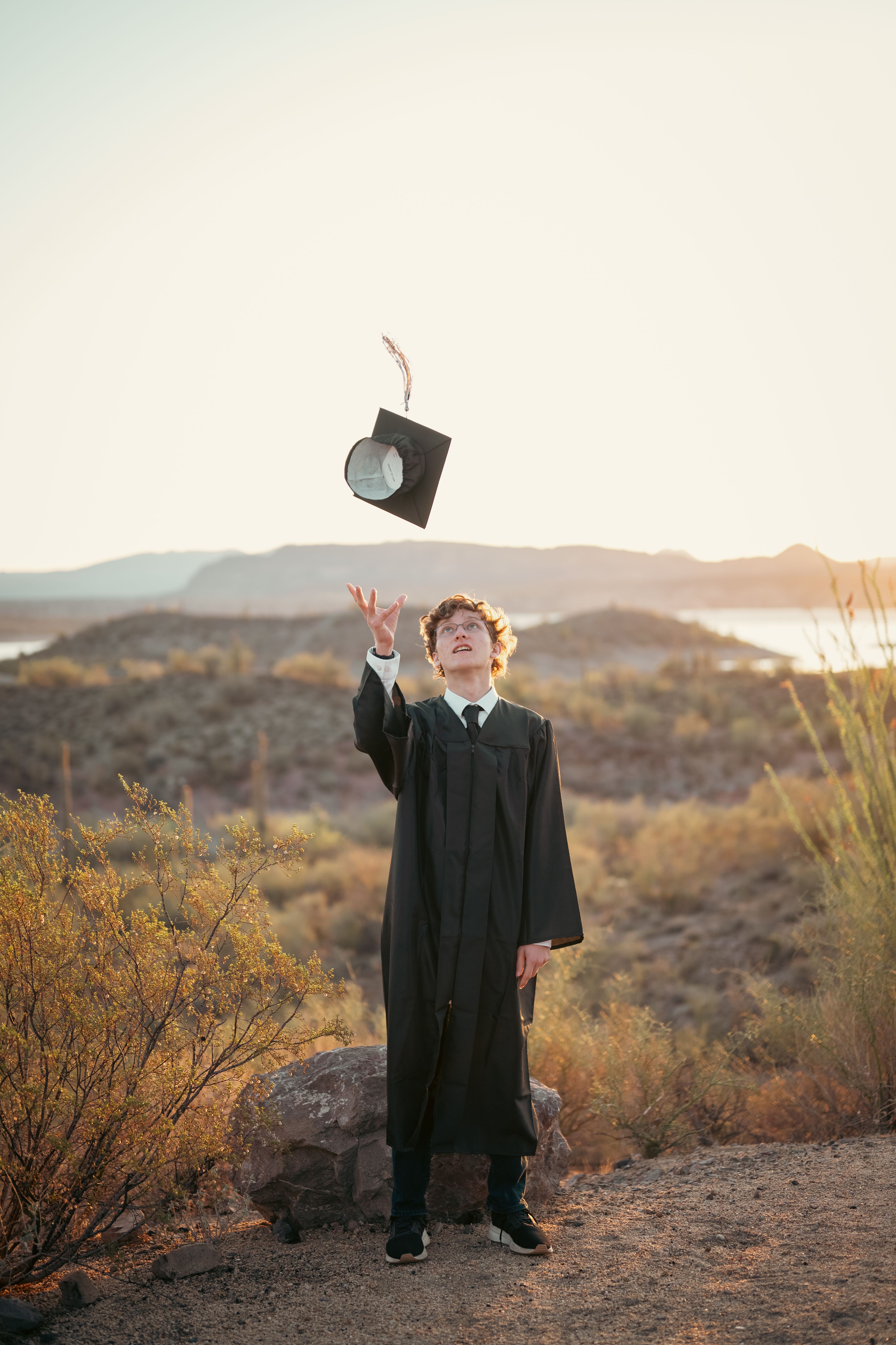 Senior photo of young man tossing graduation cap in the air with Lake Pleasant Arizona in the backdrop.