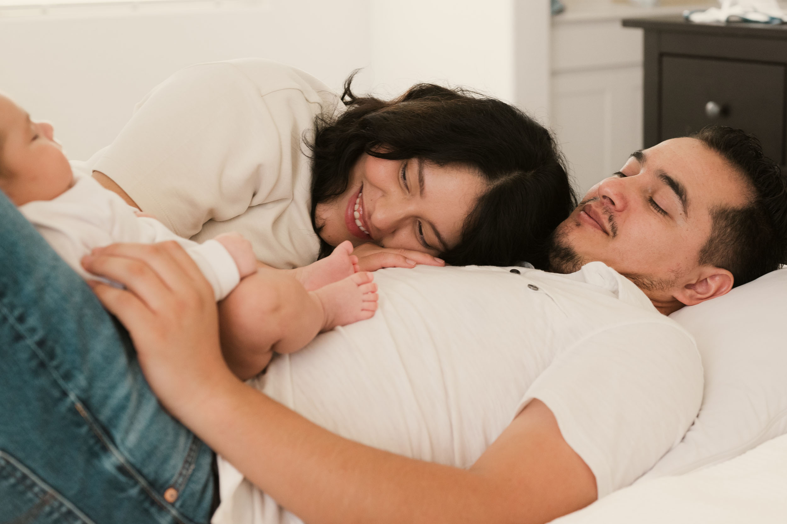 mother resting head on her husband's chest as they stare lovingly at their newborn baby