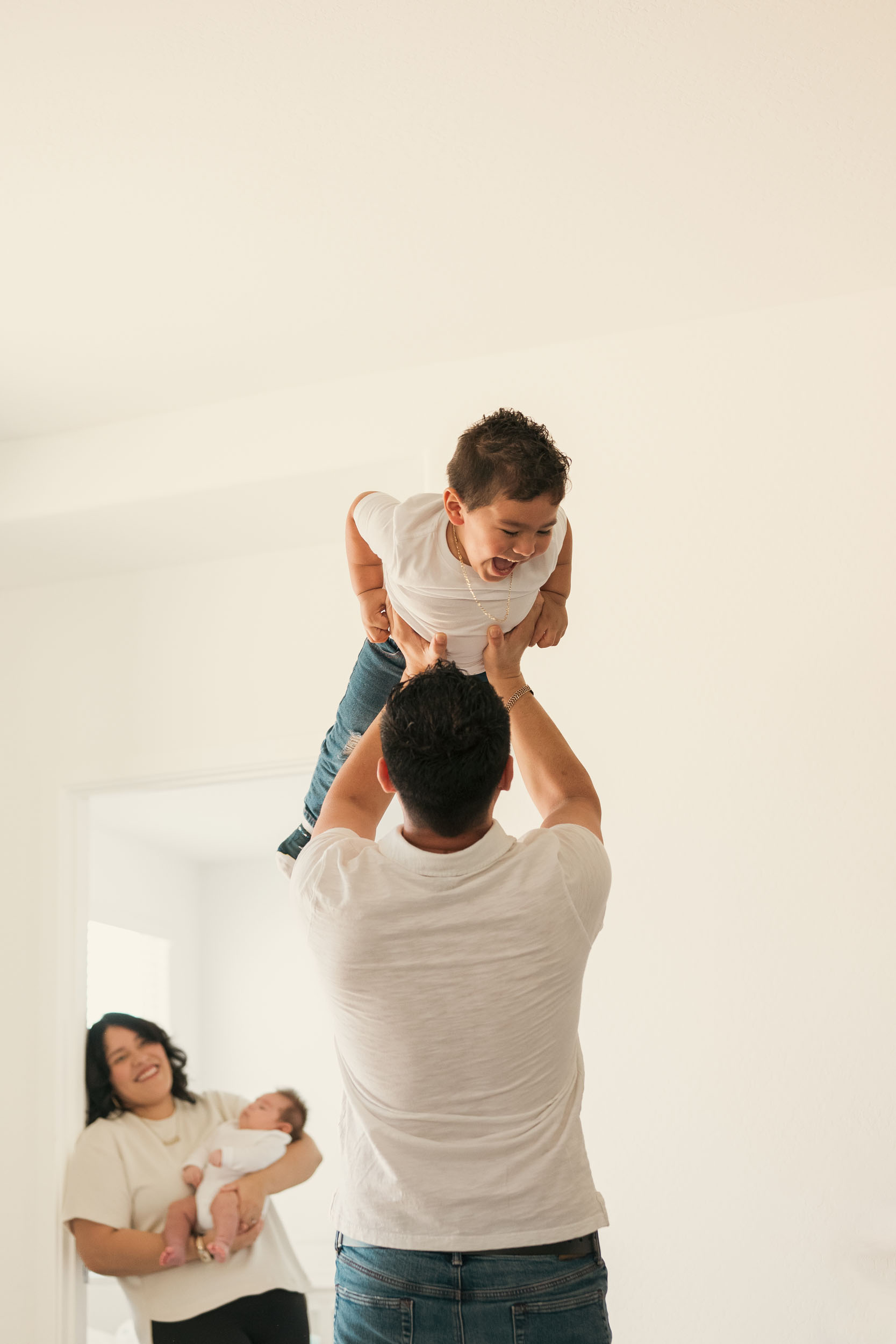 father playing with laughing young son, lifting him over his head as mother watches while holding baby