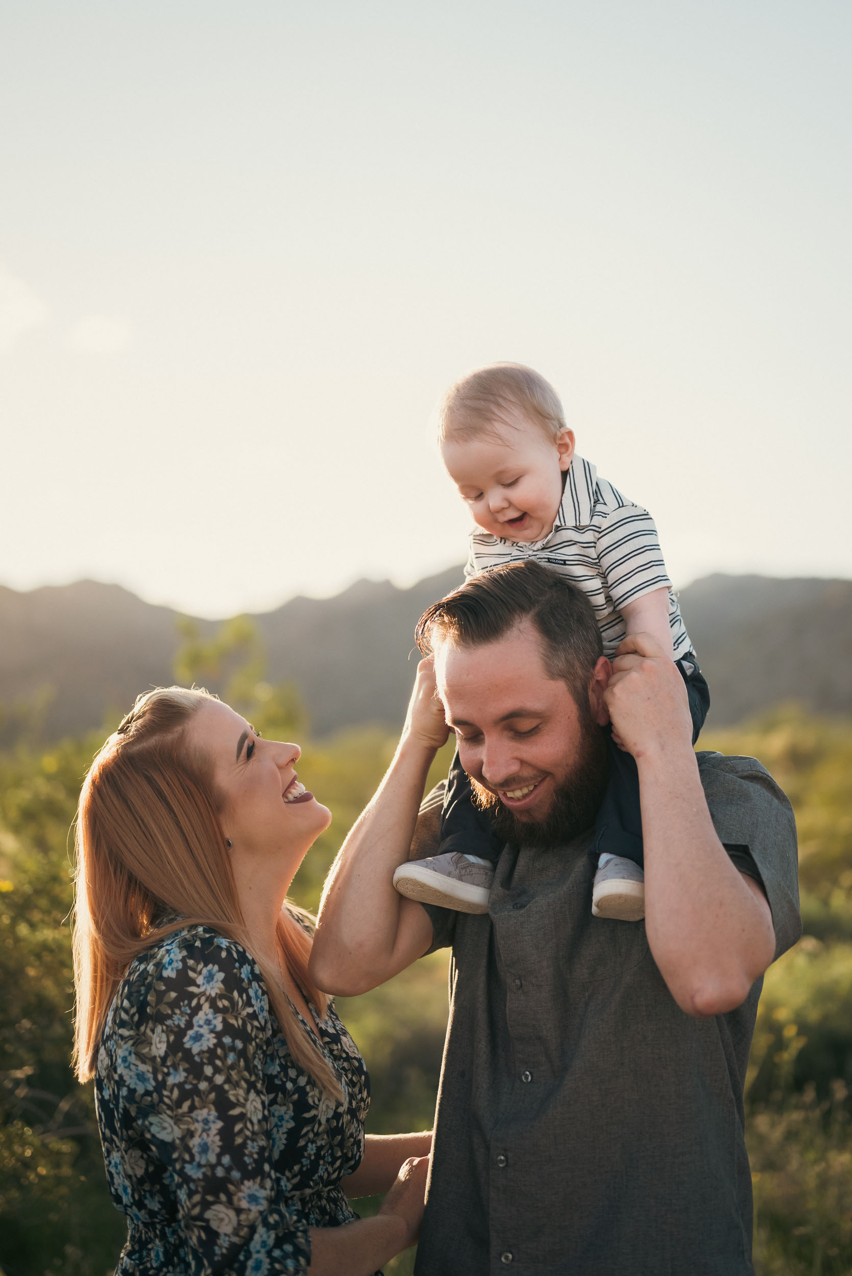 baby sitting on father's shoulders while mother smiles and admires her son