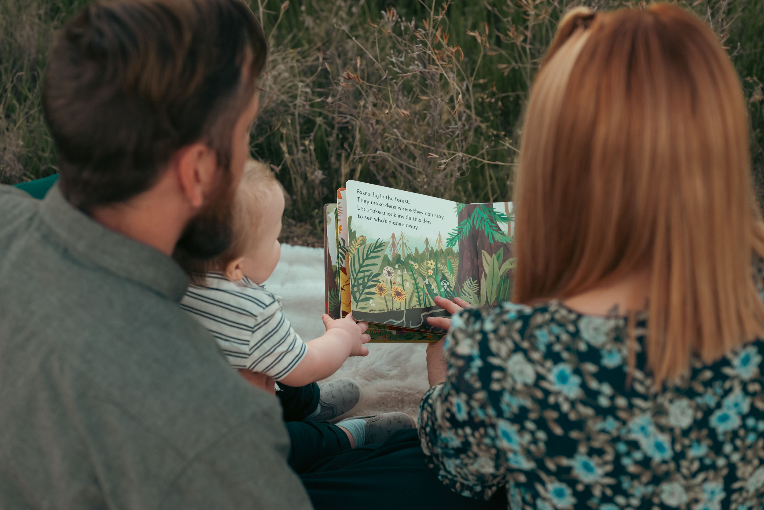 mother and father reading illustrated children's book to baby son while sitting on a blanket in nature