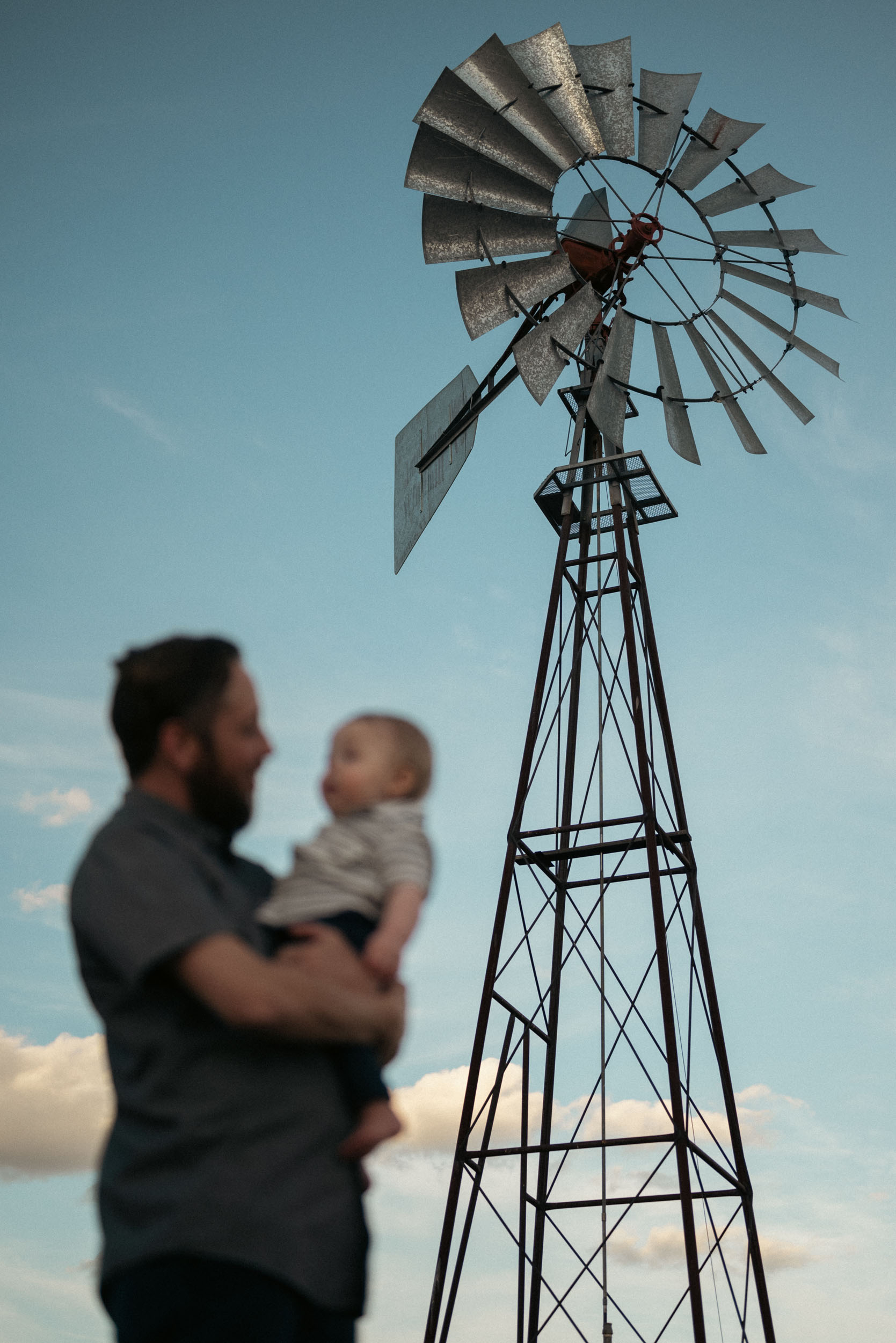 father holding baby son in foreground with tall windmill in background