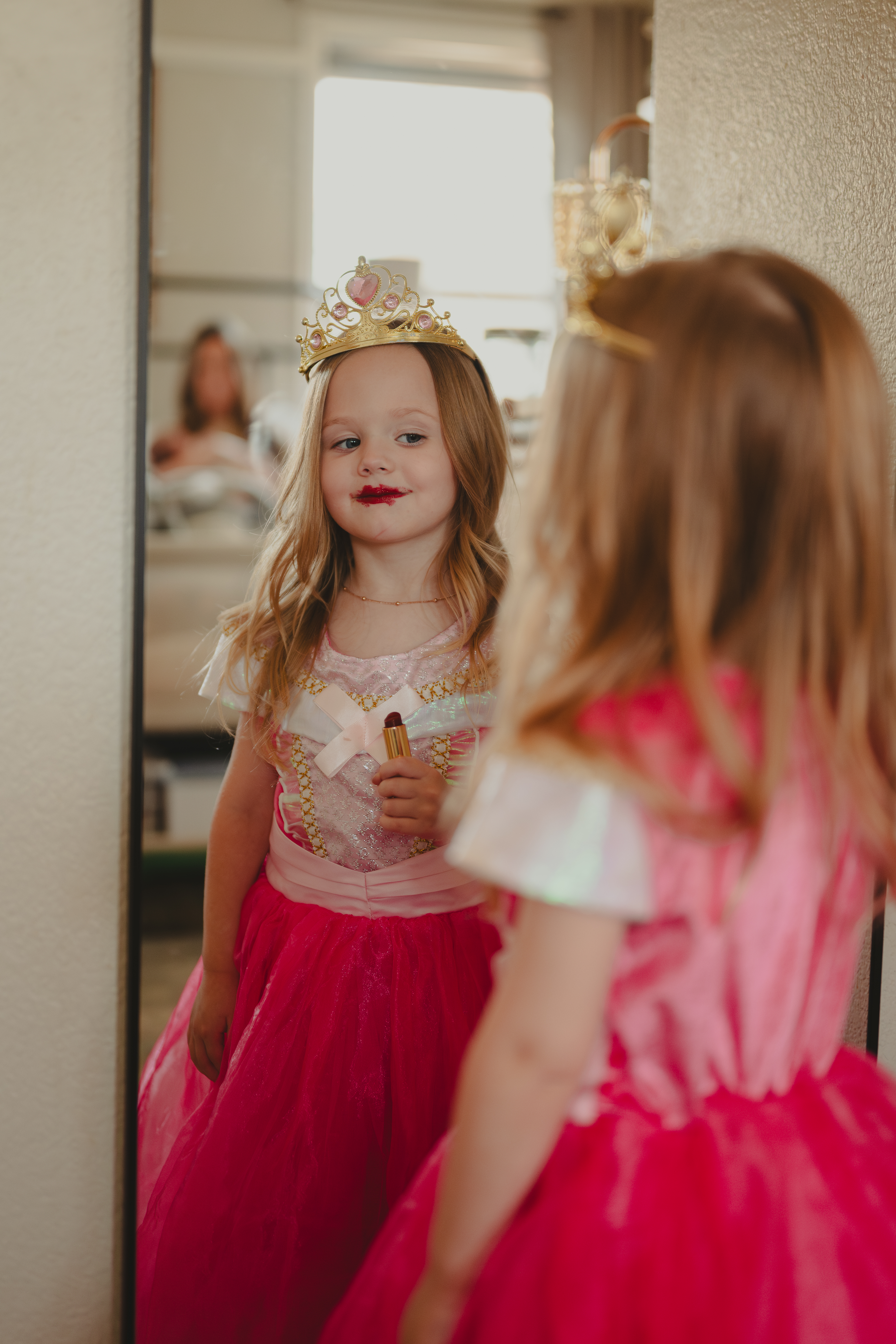 Young girl in princess costume playing with lipstick while admiring her reflection in the mirror.