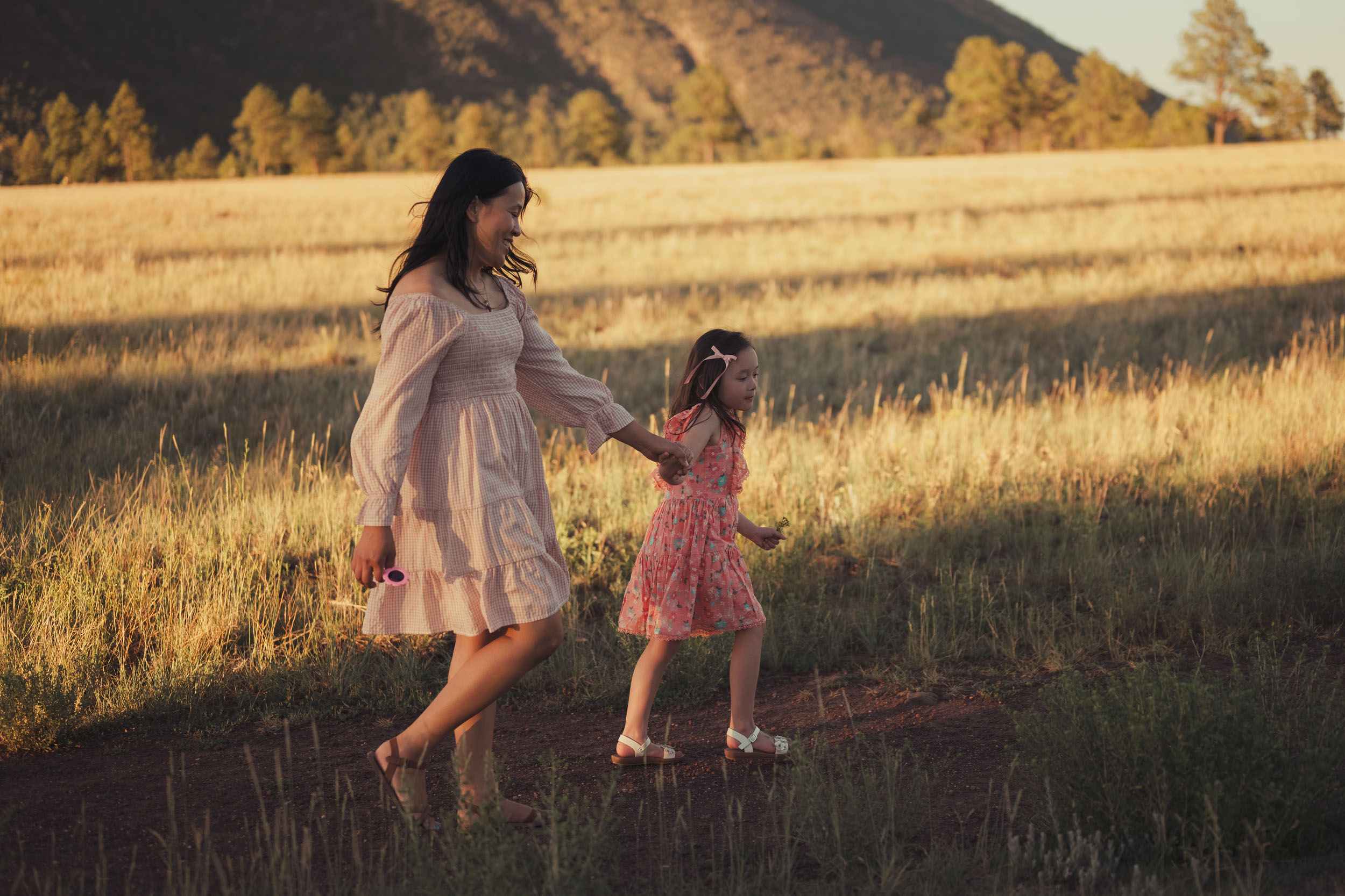 mother and daughter holding hands walking through grassy mountain meadow with golden rays of light and shadow in the background