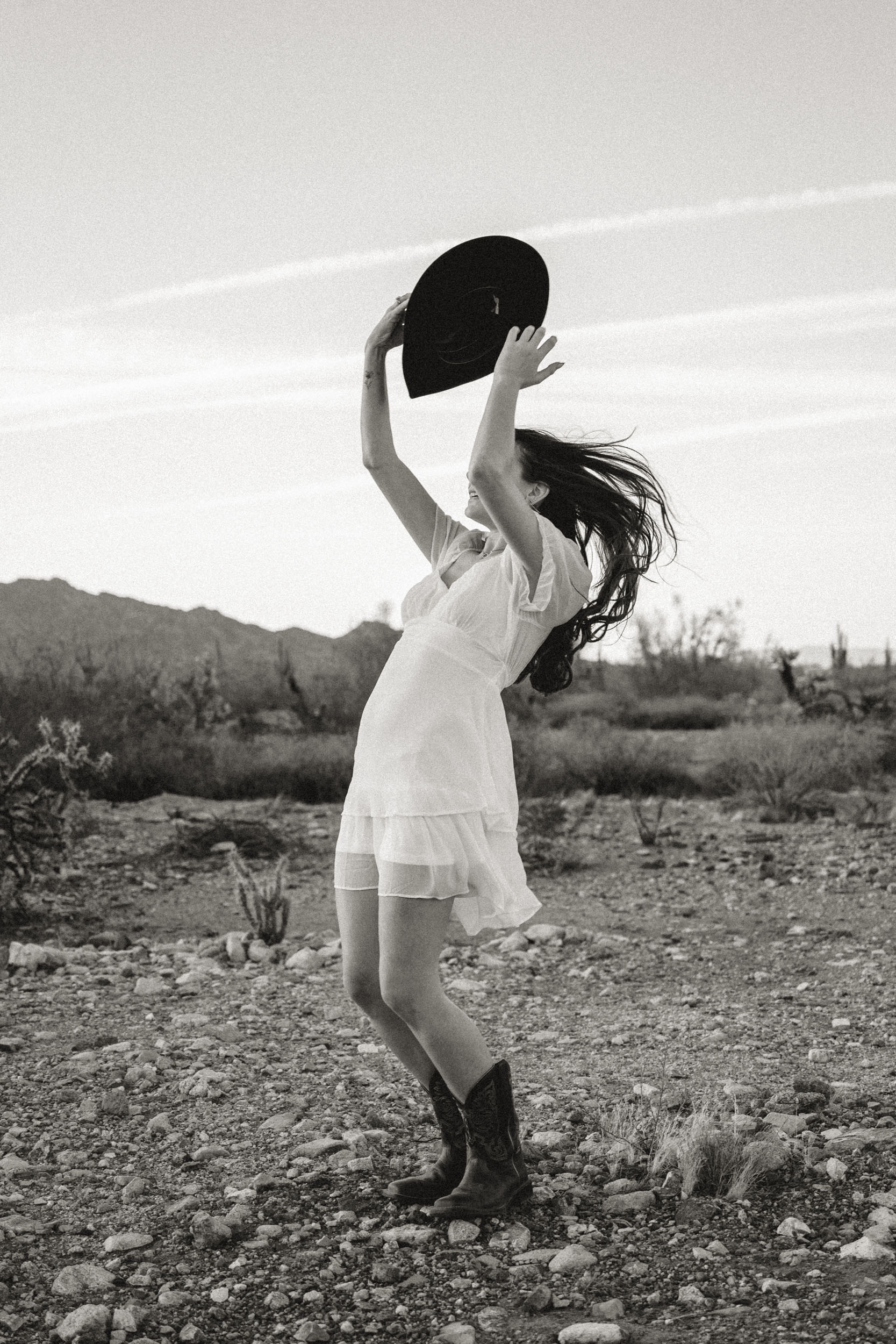 Cinematic photo of woman in dress and cowboy boots raising her hat towards the wind.