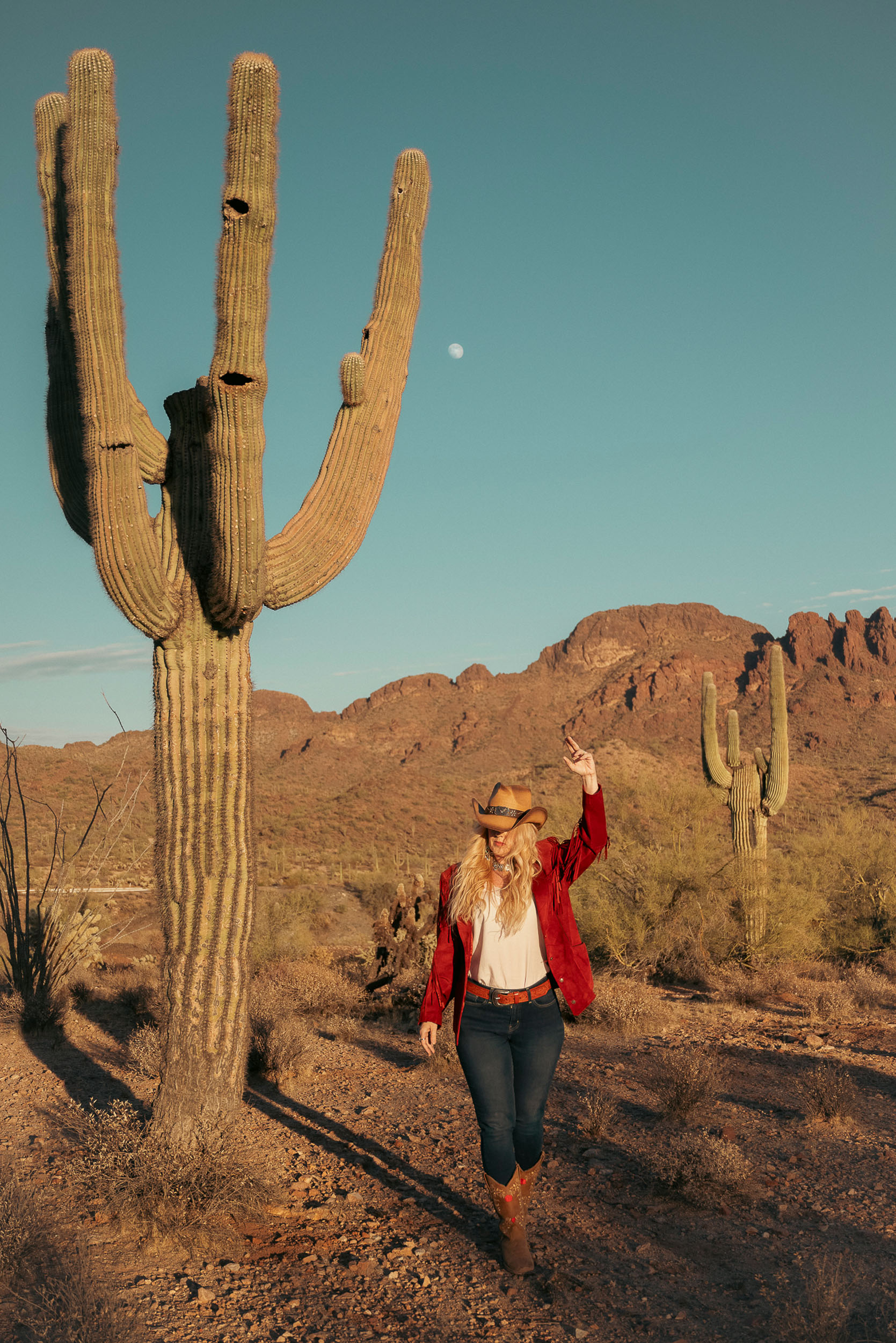 Cowgirl walking in the desert surrounded by Saguaro with Vulture Peak and the moon in the backdrop.
