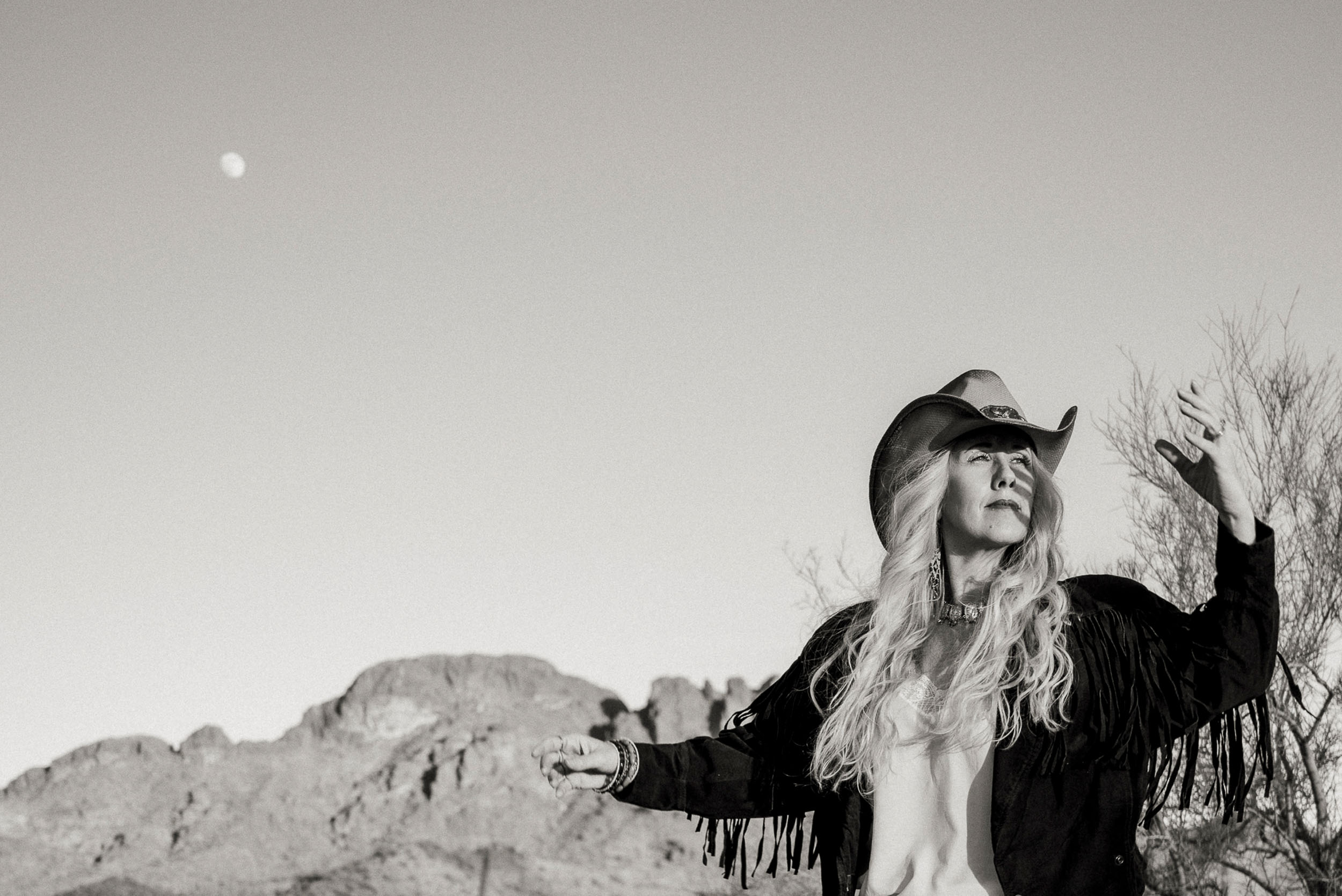 Woman in a fringed jacket and cowboy hat looking toward the sun with the moon and Vulture Peak in the backdrop.