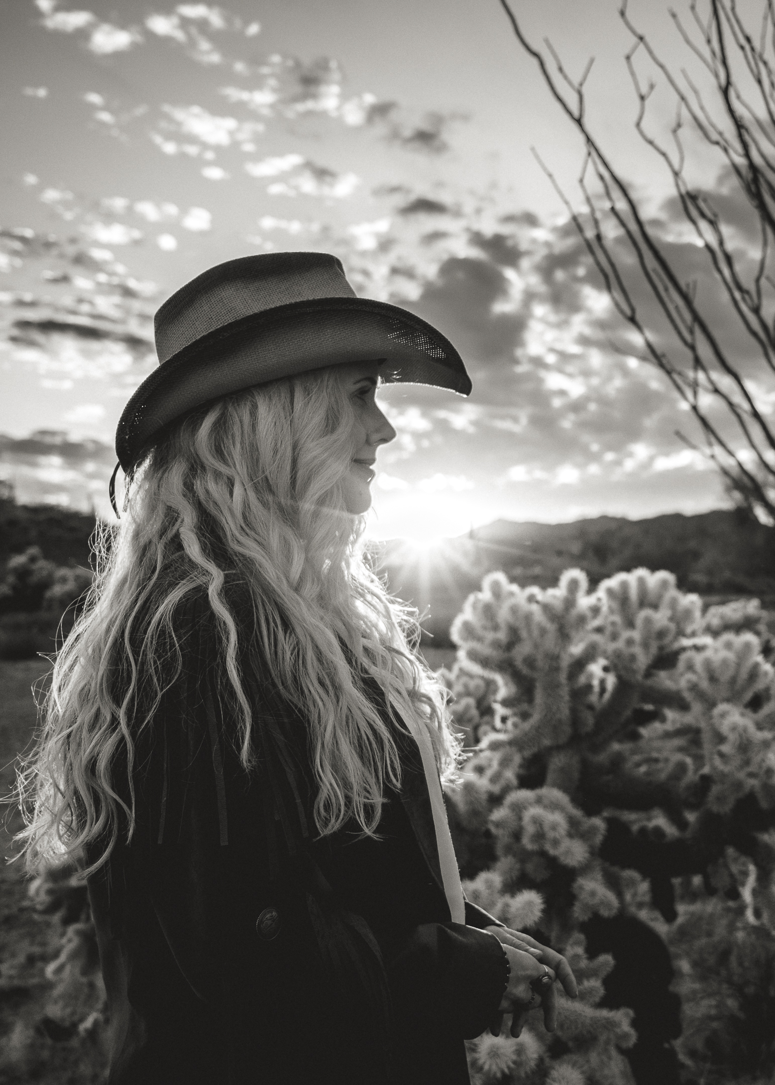 black and white photo of woman in a cowboy hat staring into the distance surrounded by desert fauna with sun setting in the background.