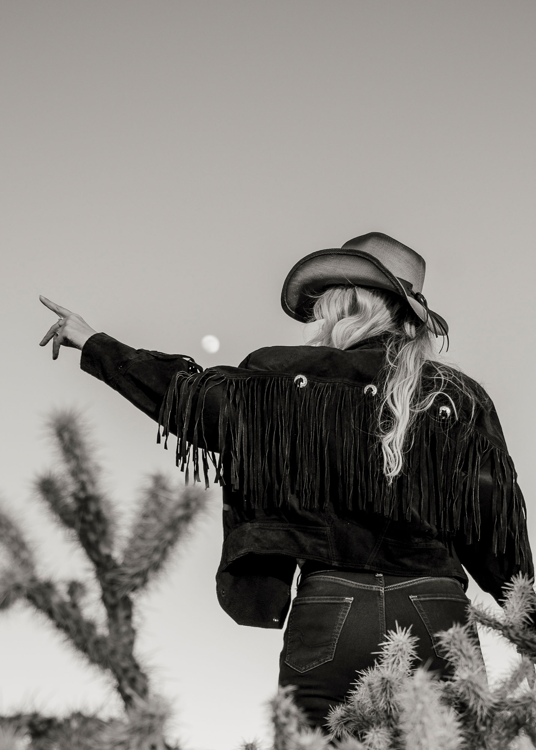 woman wearing a fringed jacket and cowboy hat pointing to the sky with the moon.